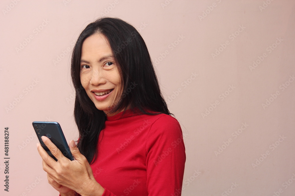 Portrait of an Asian model holding a mobile phone, turning to look at him with a smile. Wear a red sweater against a light brown background. Portrait of an Asian model holding a mobile phone, turning to look at him with a smile. Wear a red sweater against a light brown background.