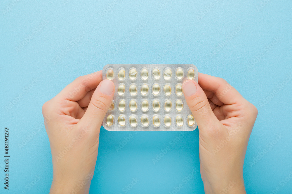 Young adult woman hands holding blister package of fish oil capsules or d vitamins on light blue table background. Closeup. Pastel color. Daily receiving nutritional supplements. Top down view. - obrazy, fototapety, plakaty 