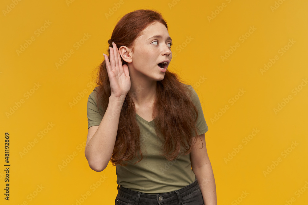 Indoor portrait of young ginger female turn her head and trying to hear something isolated over yellow background Indoor portrait of young ginger female turn her head and trying to hear something isolated over yellow background