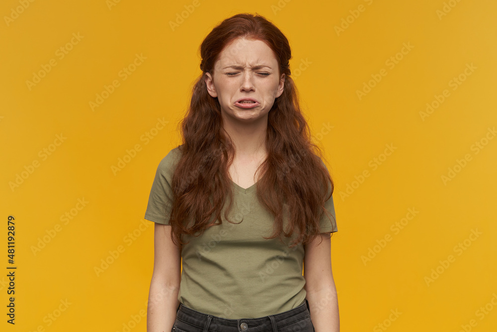 Fototapeta premium Indoor portrait of young ginger female keeps her eyes closed while crying. isolated over yellow background