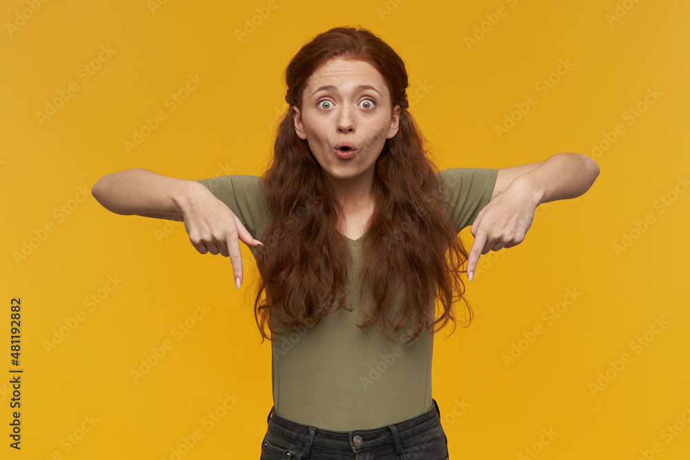 Indoor portrait of young ginger female points with a finger at bottom side, excited, shocked facial expression. isolated over yellow background Indoor portrait of young ginger female points with a finger at bottom side, excited, shocked facial expression. isolated over yellow background