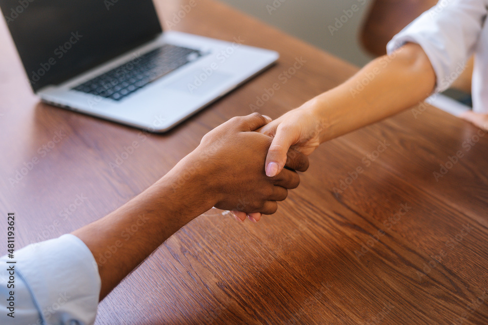 Close-up low-angle view of unrecognizable businesswoman and businessman closing deal and shaking hands in agreement. Closeup of handshake business colleagues over table with laptop, selective focus.