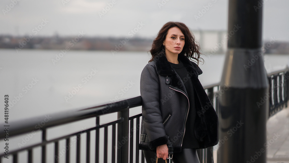 A girl in black clothes stands at the pier. Stylish look. Girl with curly hair. Against the background of the river.