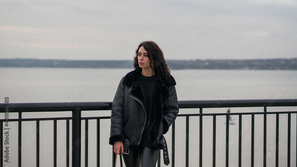 Obraz premium A girl in black clothes stands at the pier. Stylish look. Girl with curly hair. Against the background of the river.