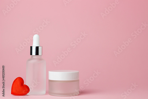 Two white glass cosmetic jars for cream and serum on a pink background with red heart. Cosmetology and beauty. Self care. Model for cosmetics.