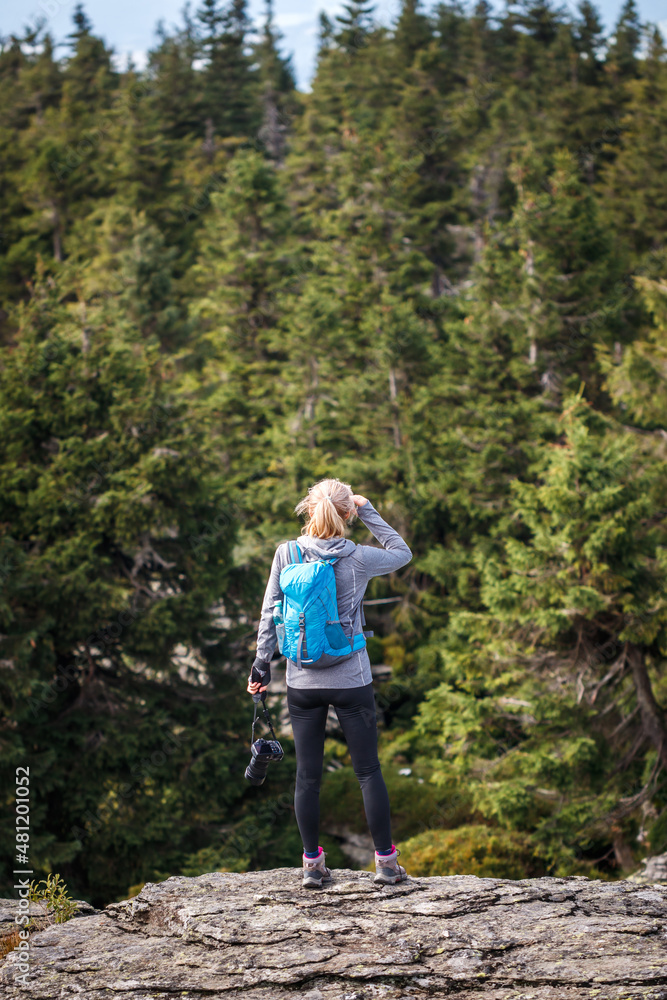 Woman hiker with camera standing at rocky mountain peak. Hiking in forest. Outdoor active lifestyle