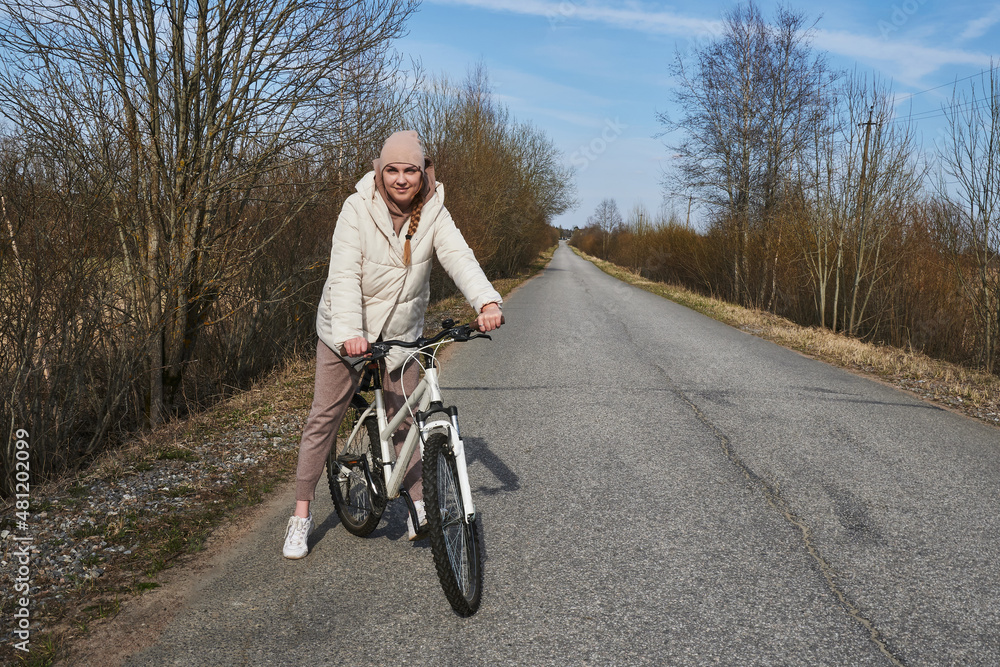young woman on a bike in a warm jacket, on a road surrounded by trees without leaves, in spring with a clear blue sky