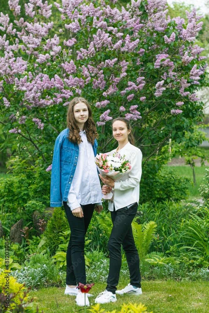 Fototapeta premium Two young beautiful girls, a portrait in the garden on a background of lilacs. Girls in a white blouse hold a beautiful bouquet of flowers in their hands.