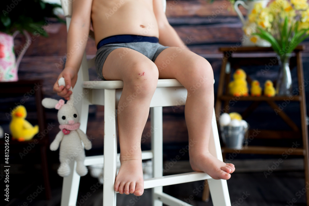 Toddler child with wounded leg, sitting on a chair, holding teddy bear rabbit