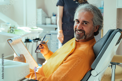 Portrait of Smiling Male Patient Filling Documents on a Clipboard while Sitting on a Dentist's Chair. Happy man looking at camera while holding anamnesis form fill and eyeglasses in dentist's office.