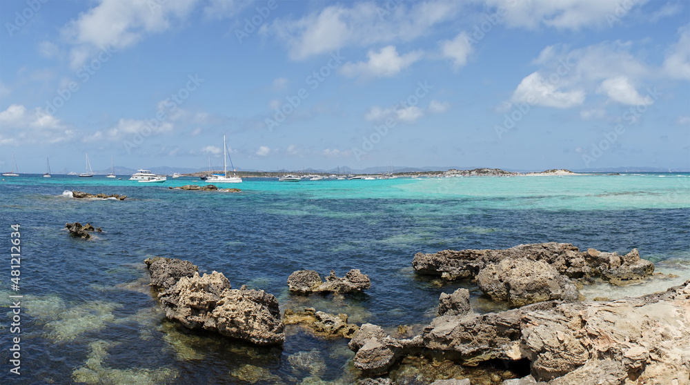 Panoramic view from a rocky beach in Formentera, Spain, in the background sailboats moored against the blue sky.