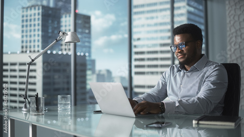 Confident Businessman in Striped Shirt Sitting at a Desk in Modern Office, Using Laptop Computer Next to Window with Big City with Skyscrapers View. Successful Finance Manager Planning Work Projects.