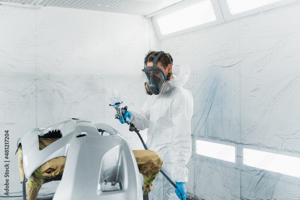 Car painter in a protective suit and mask varnishes a painted bumper of a vehicle while working in a painting booth