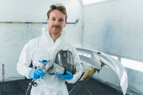 Fotografía Young car paint service worker stands in a automobile painting booth with a gun and a protective mask in his hands
