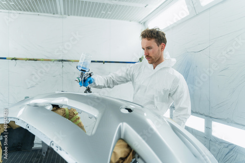 Young curly car painter varnishes a car bumper. Man works in a spray booth. Process of painting car body parts © Andrii 