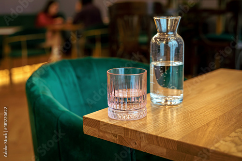 A pink glass of water and a bottle on the edge of a wooden table at the cafe, a green chair, people in the background, lifestyle image photo