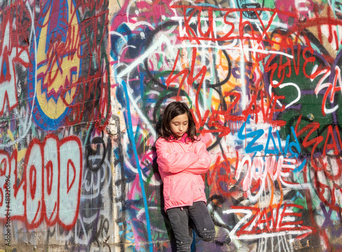 Moody pensive emotional young girl teenager with long dark hair in a pink jacket standing in front of a graffiti wall.