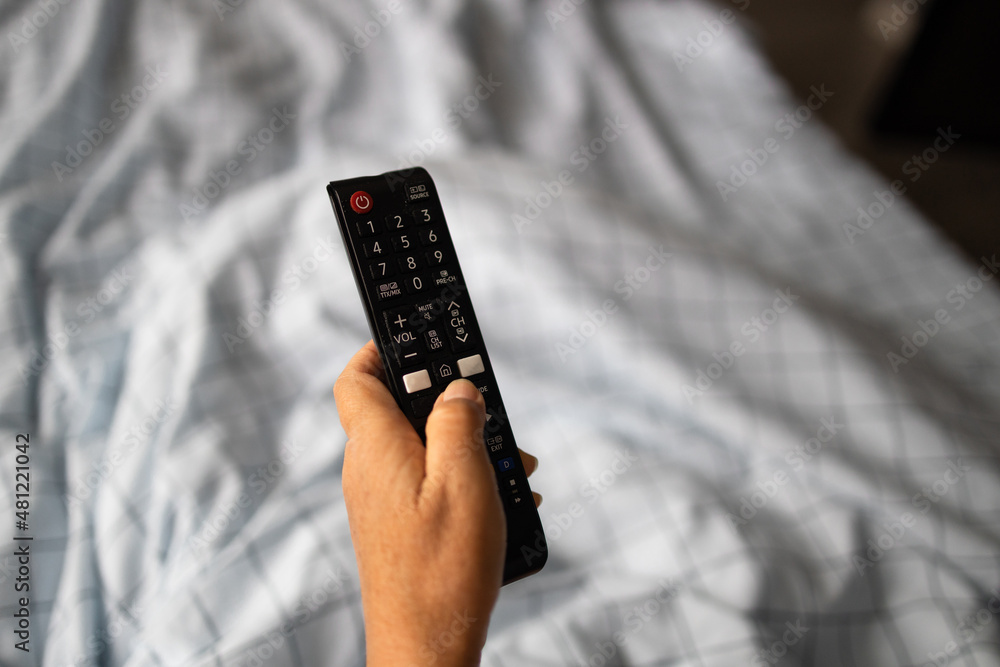 Woman hand with remote controller watching tv, enjoying sunny morning in bed in a cozy room Woman hand with remote controller watching tv, enjoying sunny morning in bed in a cozy room