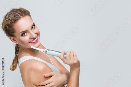 Oralcare and Dentistry Concepts. Positive Smiling Caucasian Blond Woman Cleaning Teeth with Electric Toothbrush Posing Against Over Gray Background.