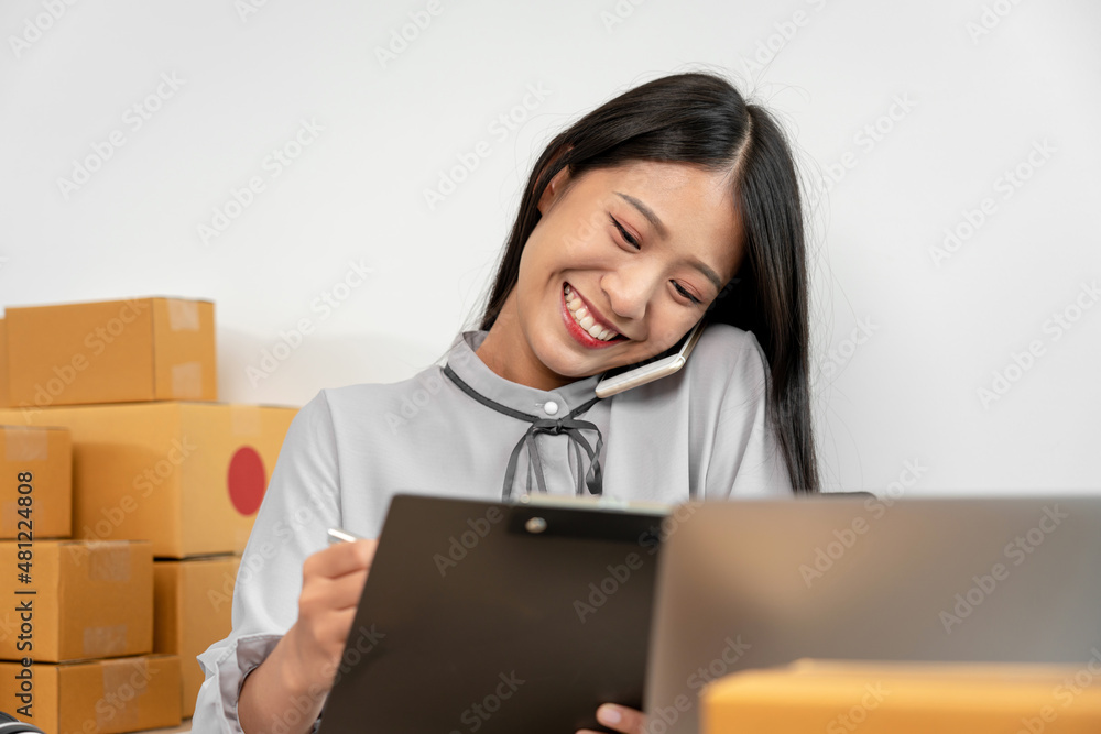 Female entrepreneur answering call from client and writing information on clipboard before packing products in parcel Female entrepreneur answering call from client and writing information on clipboard before packing products in parcel