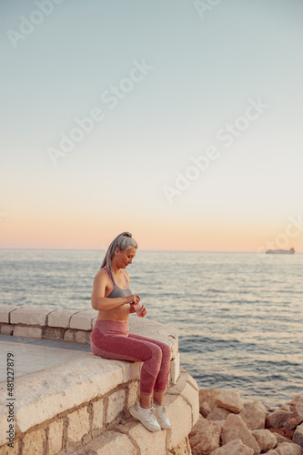 Full-length photo of asian lady in sportswear sitting on the seashore in the evening  opening a bottle of water