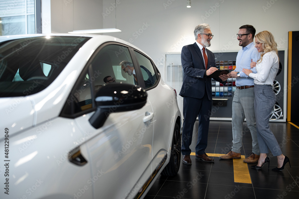 Vehicle purchase. Happy car buyers handshaking with auto seller standing among luxury automobiles in dealership showroom. Vehicle purchase. Happy car buyers handshaking with auto seller standing among luxury automobiles in dealership showroom.