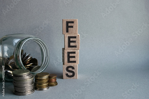 The inscription FEES on wooden cubes isolated on a light background along with a glass jar with coins, business and finance concept.