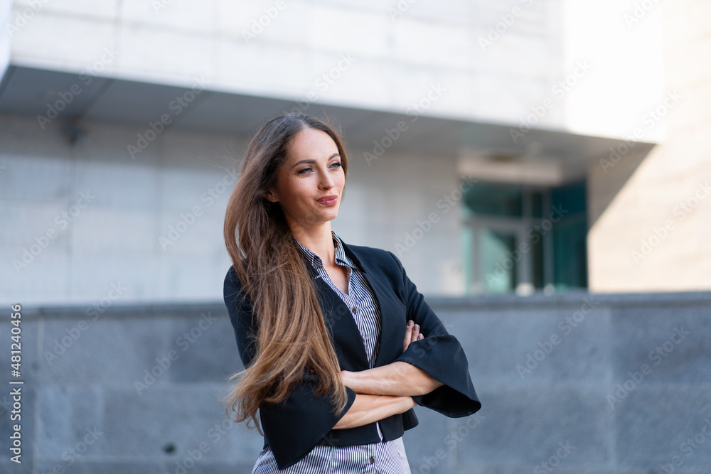 Business woman with long hair dressed black jacket standing outdoor near corporate office building hands folded