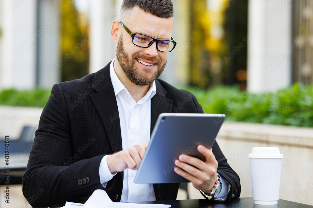 Handsome businessman in glasses sitting with tablet and cup of takeaway coffee outside on terrace Handsome businessman in glasses sitting with tablet and cup of takeaway coffee outside on terrace