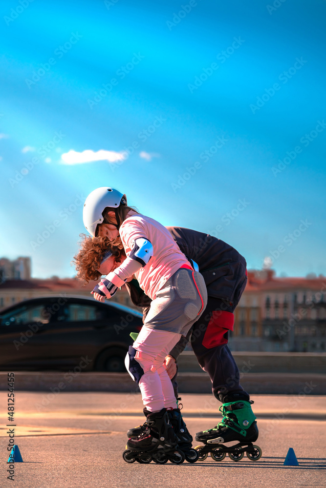 The father teaches his daughter to roller-skate in the city. The coach plays sports with the child. A girl in a helmet and fall protection is rollerblading with a man through the streets.