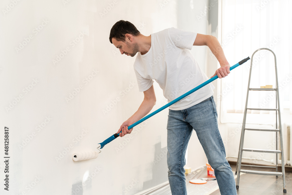 Side view of caucasian man holding paint roller and looking at the wall while painting and preparing his apartments Side view of caucasian man holding paint roller and looking at the wall while painting and preparing his apartments