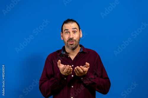 Latino man with a beard wearing a purple shirt offering with both hands isolated on blue studio background. Hospitality concept.