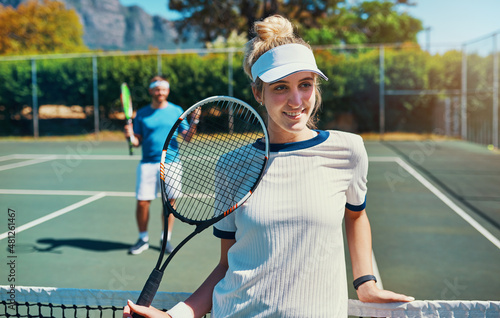 I'm always on the winning side. Cropped shot of an attractive young female tennis player outdoors on the court with her male teammate in the background. © Allistair F/peopleimages.com