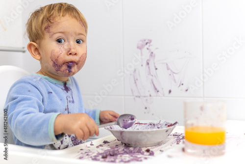 Niño rubio con una expresion divertida y cara manchada es cogido infraganti ensuciando la mesa de la cocina con la comida del desayuno