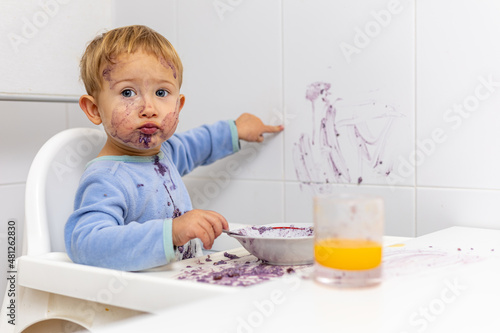Niño rubio con la cara manchada señala la pared que acaba de manchar con su comida