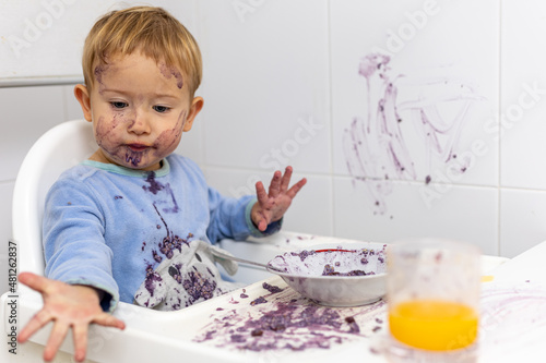 Un niño rubio de 2 años con pijama azul sentado en una trona con la cara sucia muestra la mano después de ensuciar la mesa y la pared con la comida de su desayuno.
