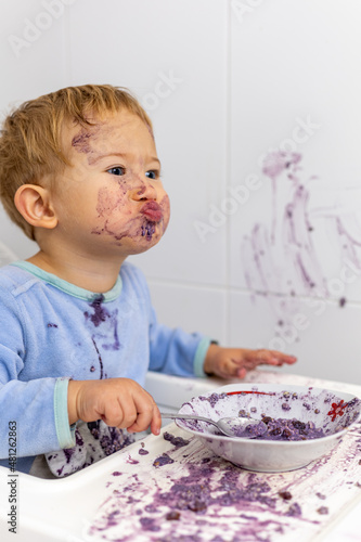 Foto en vertical de un niño rubio con pijama azul con expresión graciosa de sus labios con toda la cara manchada de arándanos de su desayuno. La mesa y la pared estan tambien manchadas