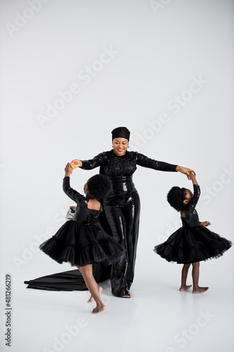 African muslim woman in elegant black outfit and hat holding hands with her graceful little daughters in stylish dresses over white background. Proud of being mother of two female kids.