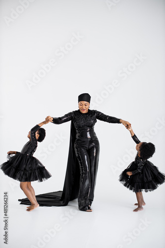 African islamic woman in elegant black outfit and hat holding hands with her graceful little daughters in stylish dresses over white background. Proud of being mother of two female kids.