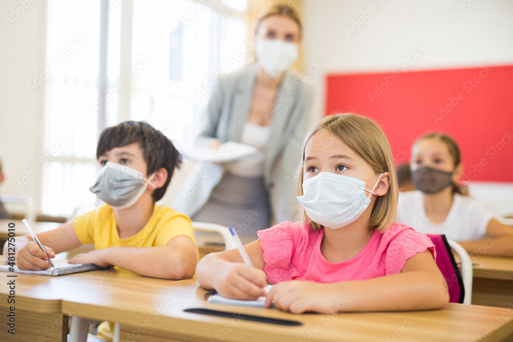 Fototapeta premium Portrait of cute little schoolgirl in protective face mask studying in classroom. Necessary precautions in coronavirus pandemic..