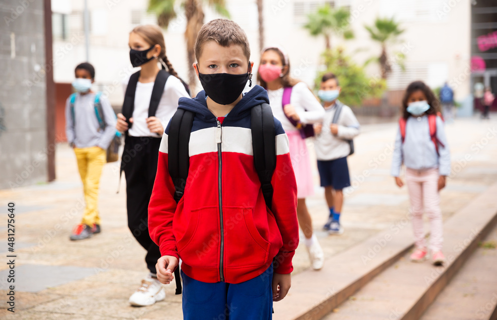 Obraz premium Confident tweenager in protective mask walking with other schoolchildren to school campus after lessons on spring day. Concept of necessary precautions in COVID pandemic