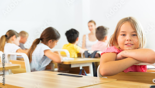 Portrait of cheerful cute blonde tween schoolgirl sitting at school desk at lesson in class, smiling looking at camera..