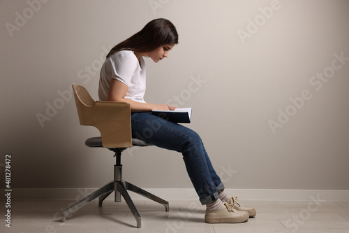 Young woman with bad posture reading book while sitting on chair near grey wall. Space for text