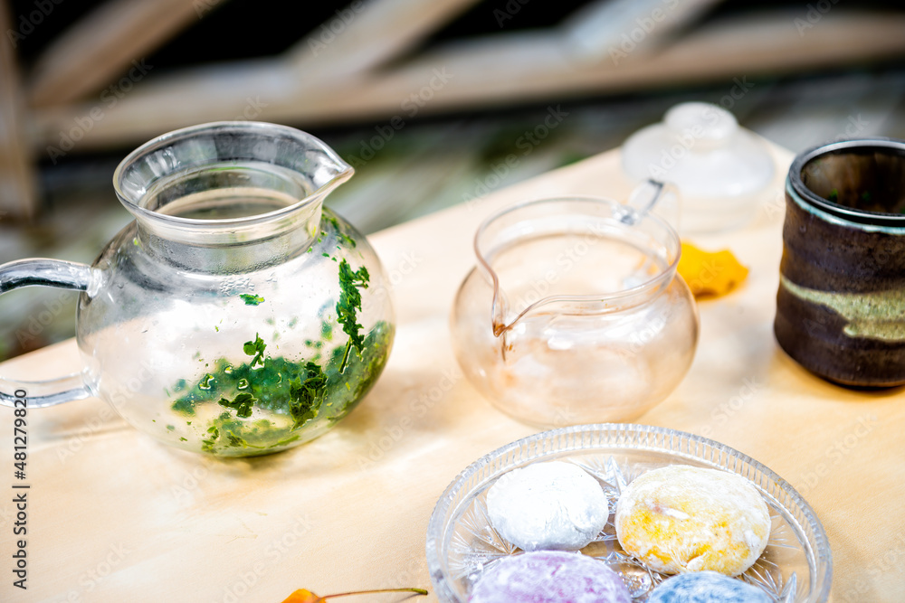 Green tea leaves in glass teapot with colorful mochi rice cake Japanese dessert daifuku on wooden table still life closeup Green tea leaves in glass teapot with colorful mochi rice cake Japanese dessert daifuku on wooden table still life closeup