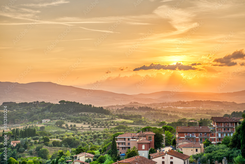 Sunset in small town village of Chiusi Tuscany Italy with houses roof rooftops on mountain countryside rolling hills landscape and orange yellow colorful evening sky with sun rays