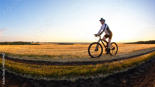 The cyclist rides a bike on the road near the field against the backdrop of the setting sun. Outdoor sports. Healthy lifestyle.