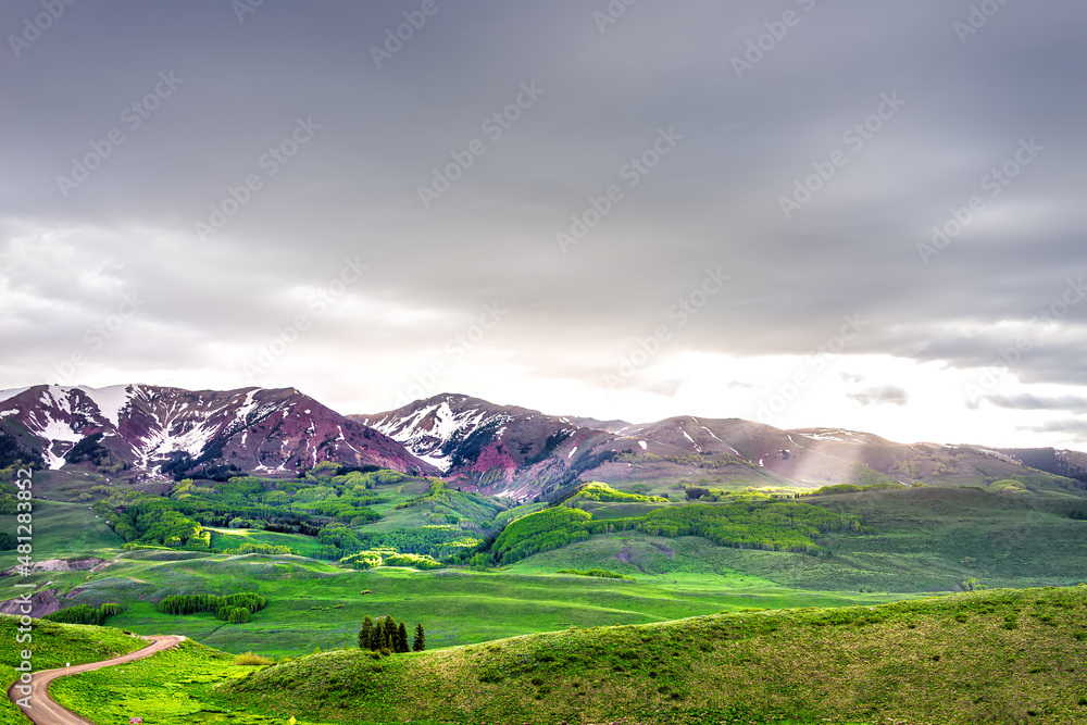 Mount Crested Butte, Colorado alpine meadow valley view from Snodgrass hiking trail in summer with green grass field and sun rays god beams through clouds with gothic rocky mountain
