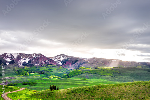Mount Crested Butte, Colorado alpine meadow valley view from Snodgrass hiking trail in summer with green grass field and sun rays god beams through clouds with gothic rocky mountain