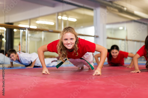 Joyful young girls and boys in karate uniform doing push-ups in gym.