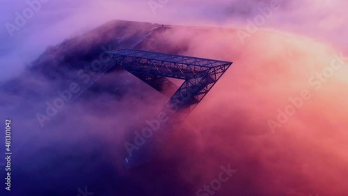 Monument on mountain in early morning light with sun shining from one side and clouds passing over the top, drone shot